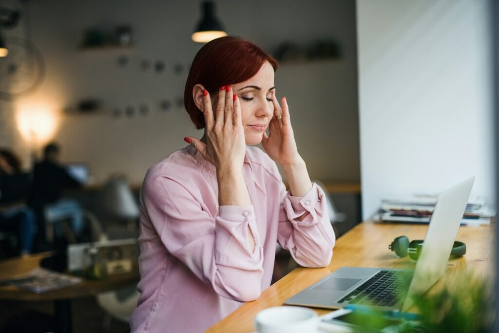 A woman sits at her laptop in a cafe massaging her head after suffering with eye strain