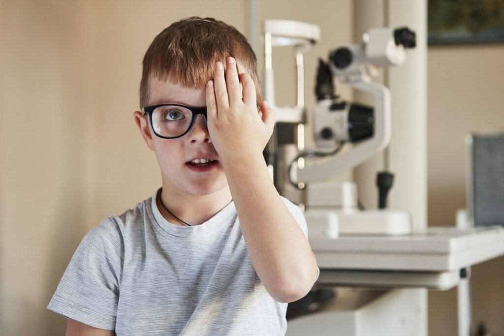 A boy with a lazy eye and glasses covers one eye and looks at wall at opticians