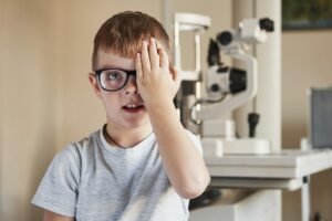 A boy with a lazy eye and glasses covers one eye and looks at wall at opticians